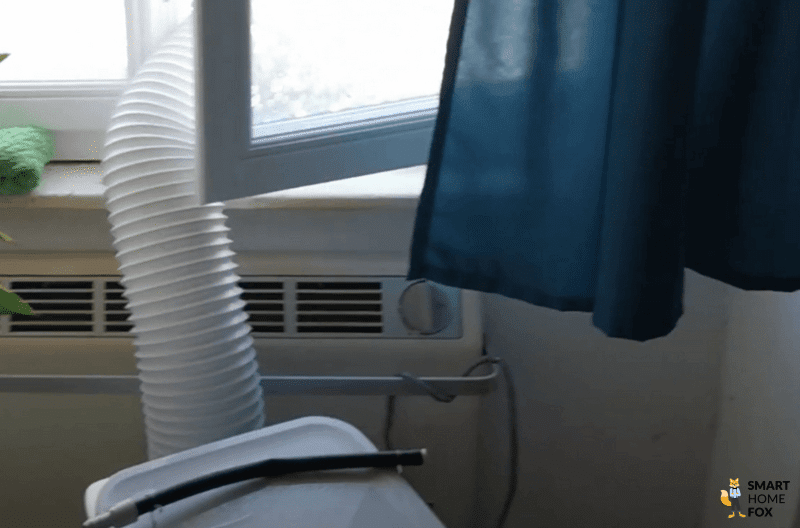 Eine mobile Klimaanlage steht vor einem Fenster und der Abluftschlauch hängt aus dem geöffneten Fenster heraus.